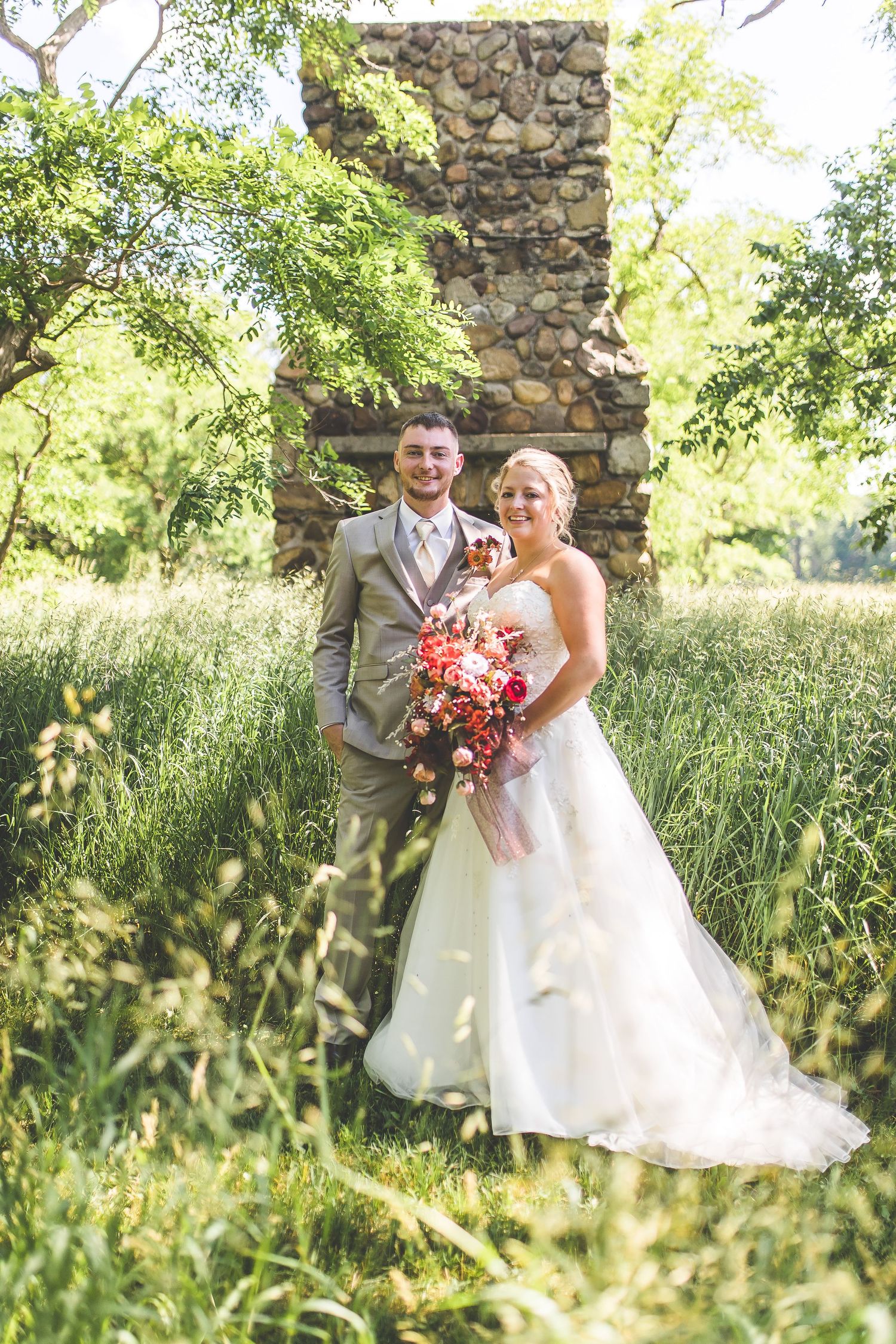 A couple poses in front of a rustic stone wall surrounded by tall summer grasses and greenery.