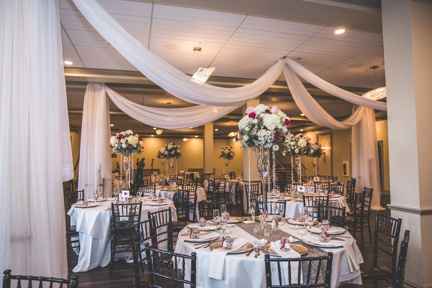 Elegant wedding reception hall decorated with white draping and dark chiavari chairs.