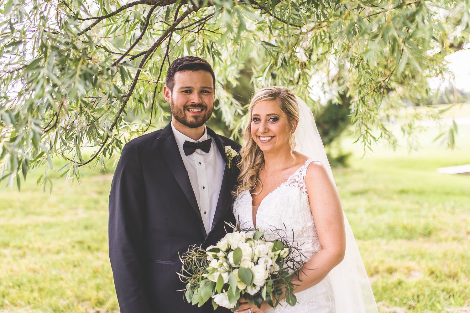 Newlyweds pose together holding a white floral bouquet under willow trees in an outdoor garden setting.