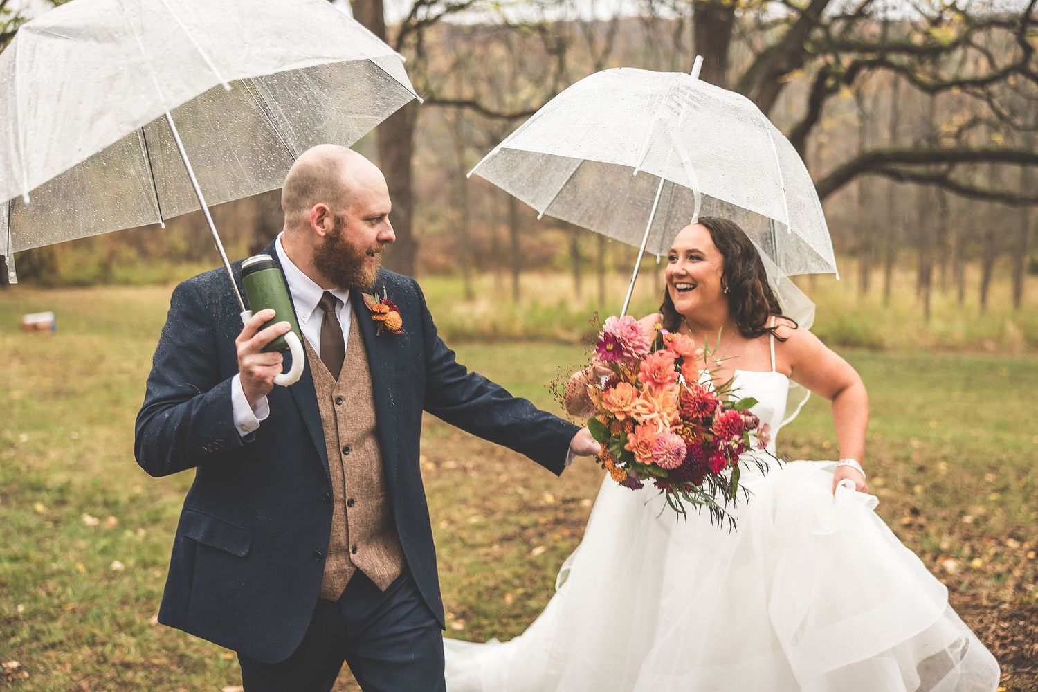 A whimsical wedding photo of a couple with clear umbrellas and fall bouquet in the rain.