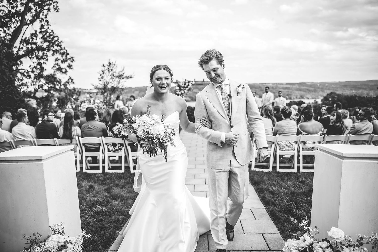 Wedding guests gather outdoors on a scenic hillside for a ceremony with white chairs arranged in rows.