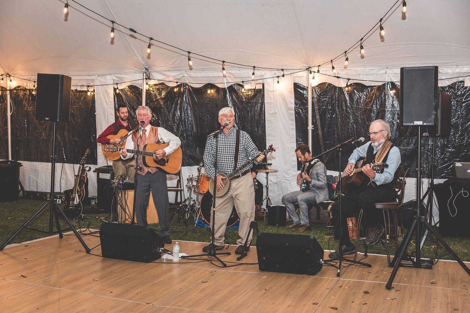 Wedding guests dance and celebrate on a wooden dance floor under warm string lights at an evening reception.