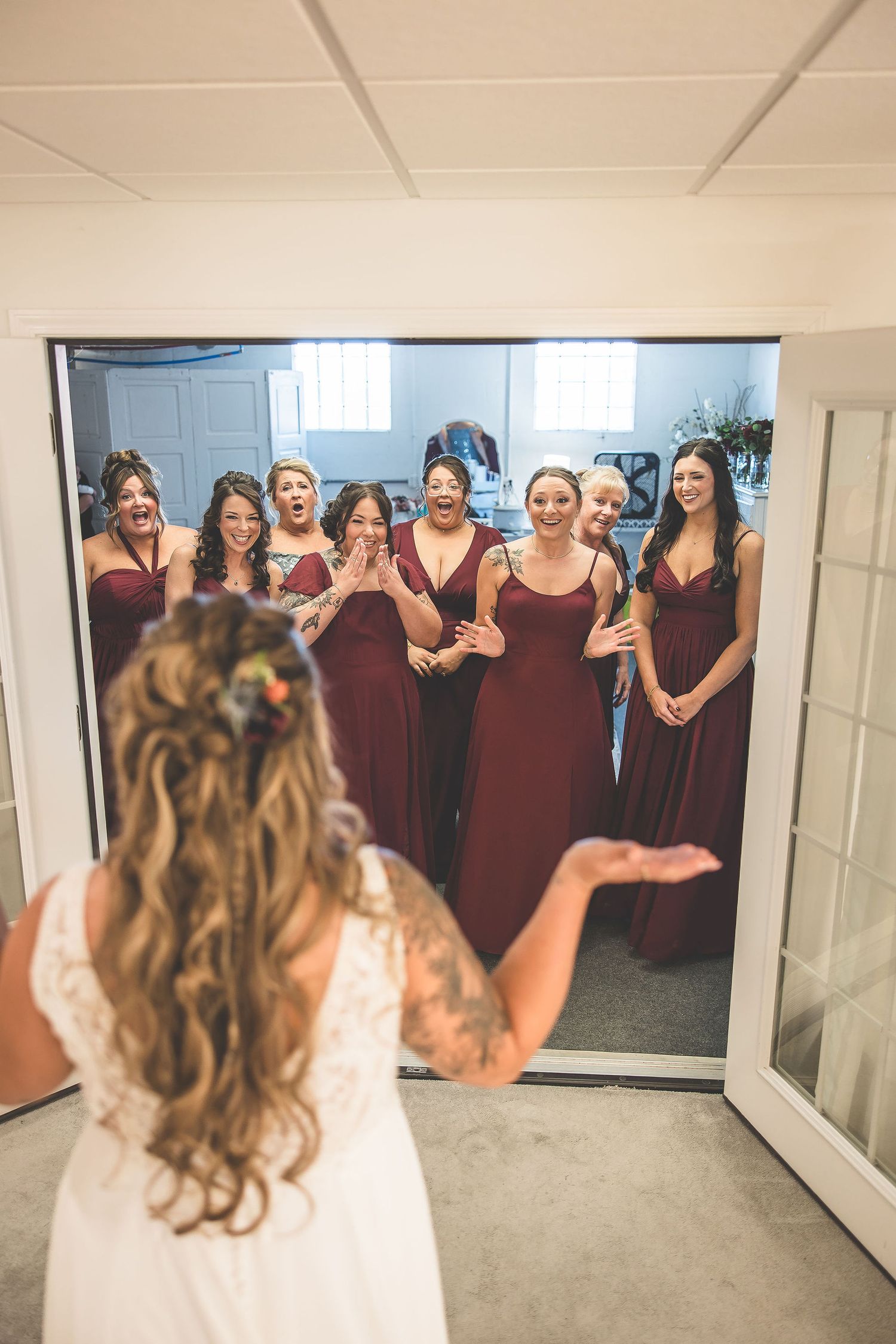 Wedding party in burgundy dresses reacts with joy as bride reveals her dress.