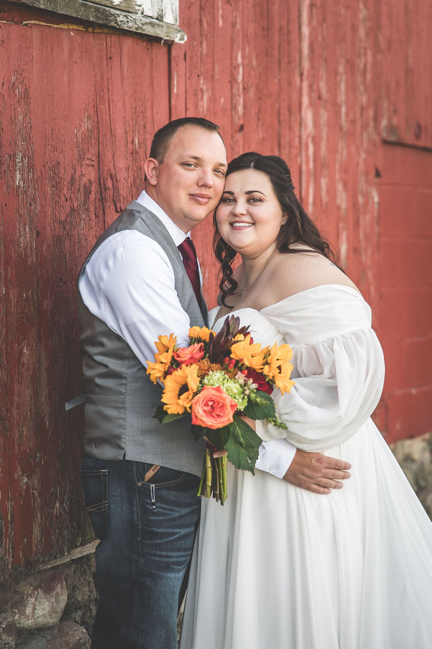 Couple with bright orange bouquet poses by rustic red barn wall.