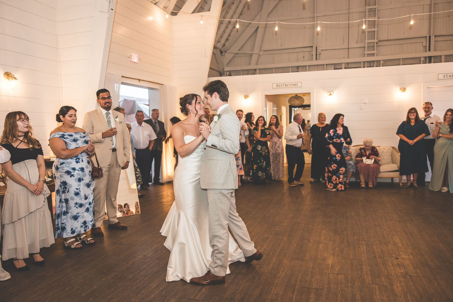 Couples dance on a wooden floor during a wedding reception in a rustic venue with string lights.