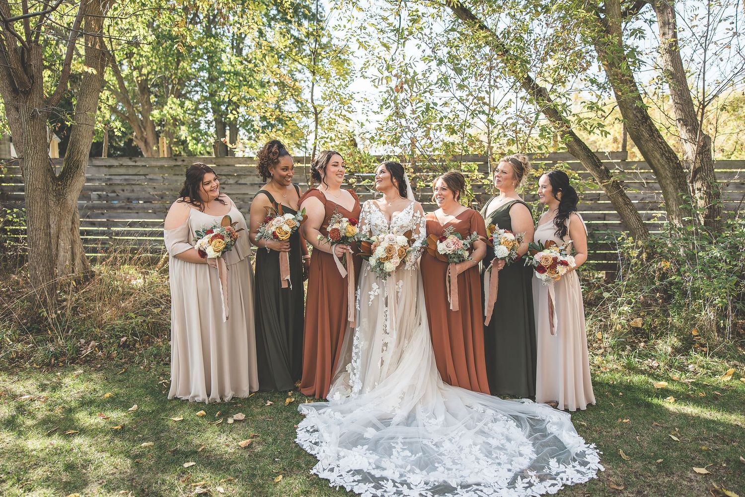 Wedding party in mix of burgundy and neutral dresses poses in natural outdoor setting.