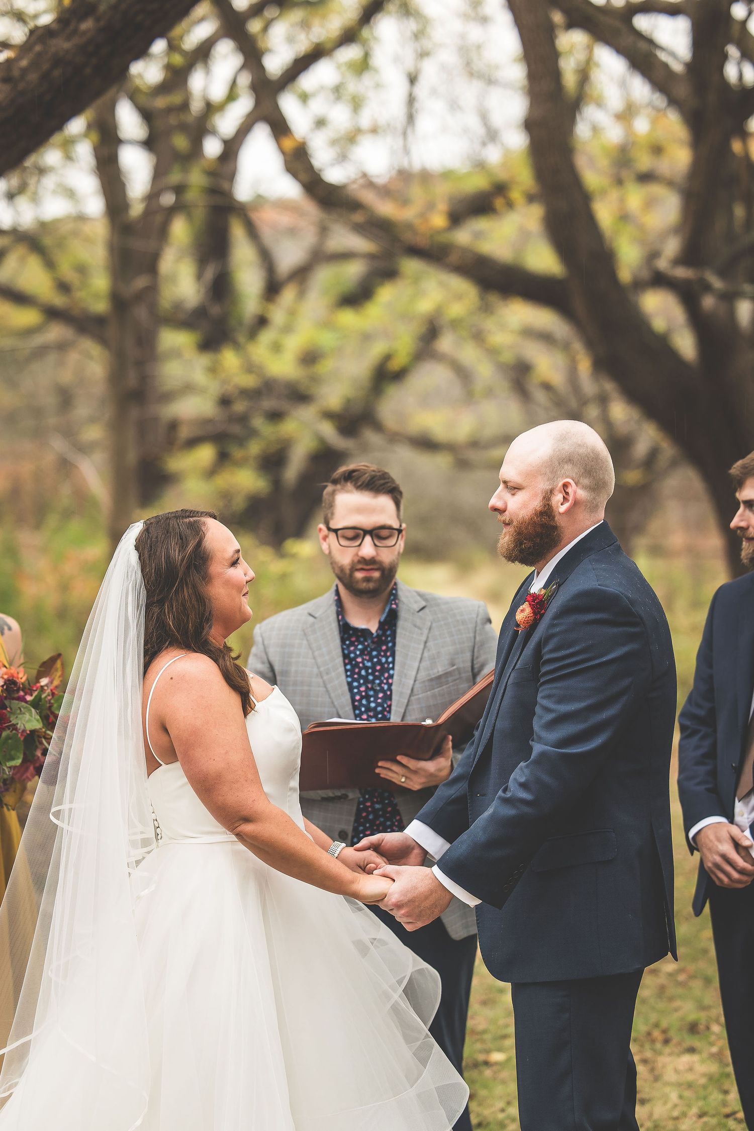 Outdoor wedding ceremony under trees with couple holding hands during vows.