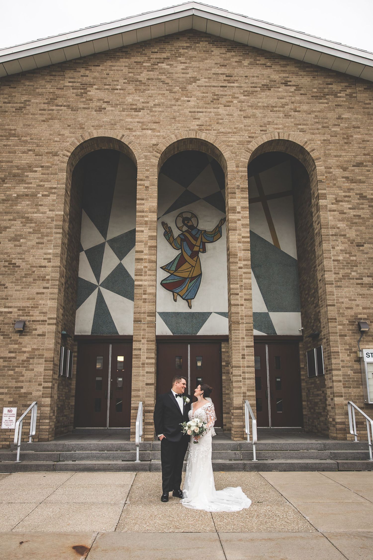 Wedding couple poses in front of modern church entrance with geometric stained glass windows.