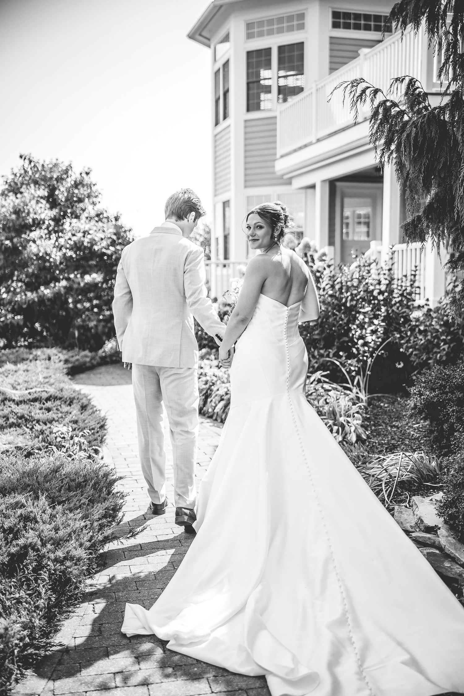 Wedding couple walks along landscaped path beside classic coastal architecture.
