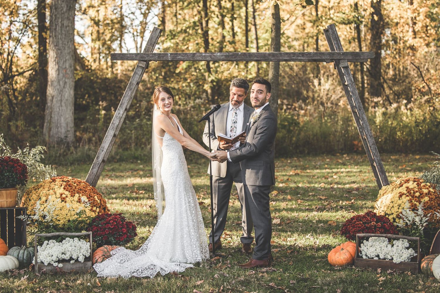 Fall wedding ceremony with couple standing under wooden arch decorated with autumn flowers.