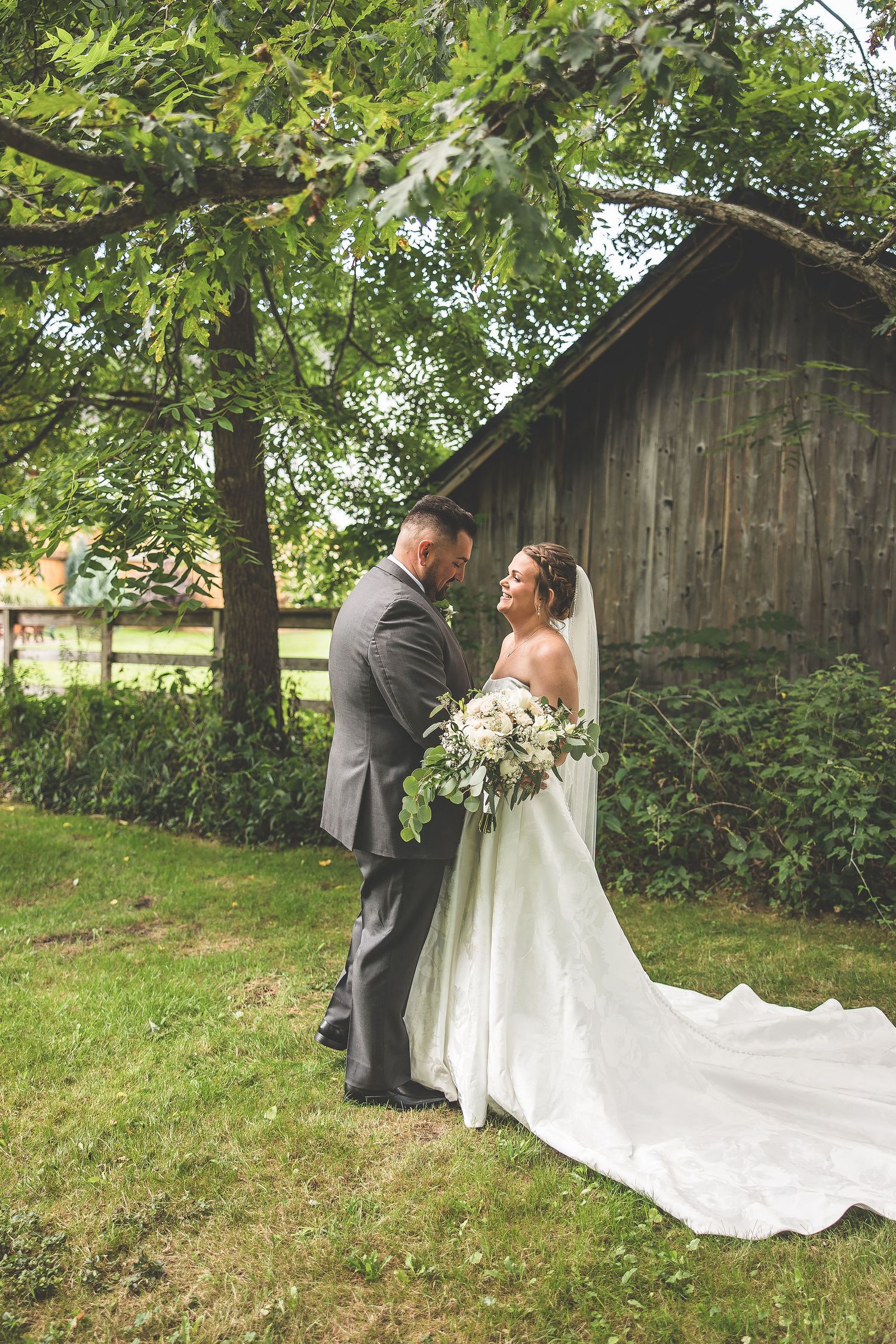 First look wedding moment unfolds near rustic barn surrounded by lush summer greenery.