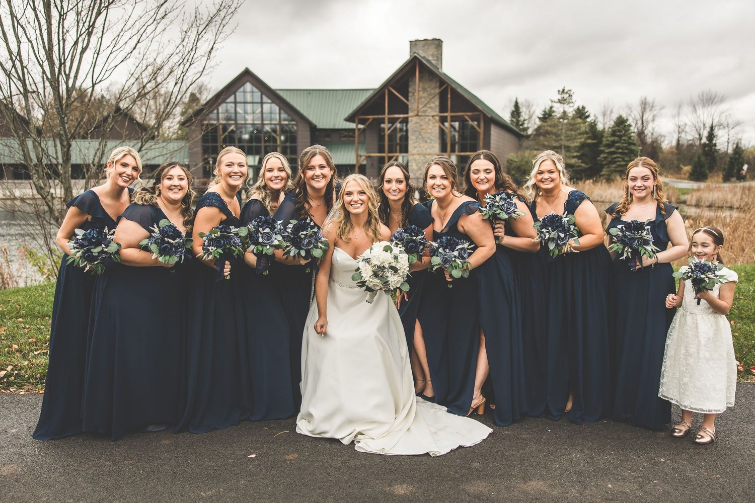 Wedding party members gather outdoors near rustic building for formal group photos.
