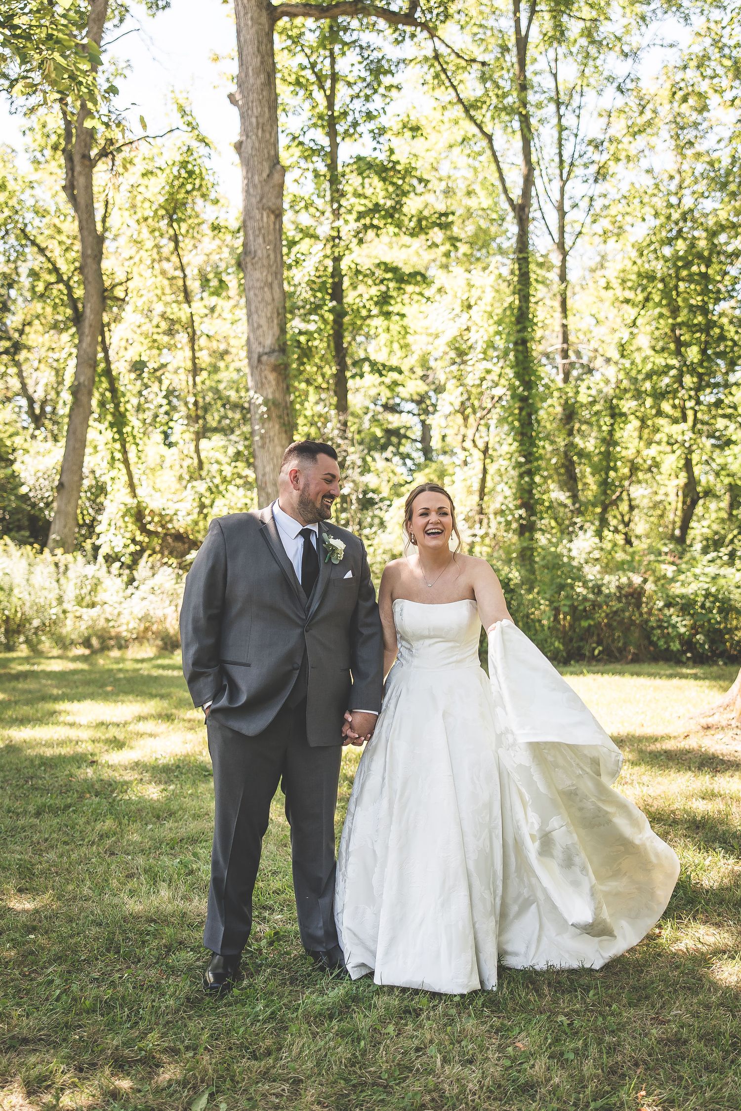 Candid wedding portrait of couple walking and laughing in sunlit woodland setting.