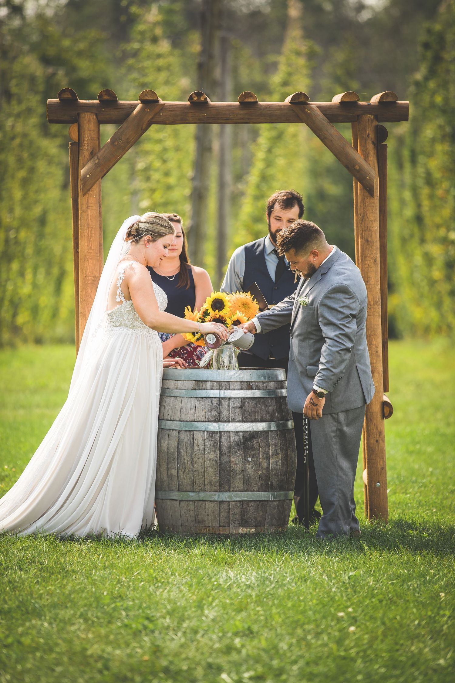 A rustic outdoor wedding ceremony featuring a wooden barrel altar and sunflowers in a lush green field.
