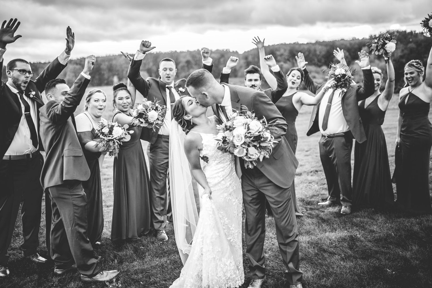 Wedding guests form a joyful line outdoors on a grassy field under cloudy skies.