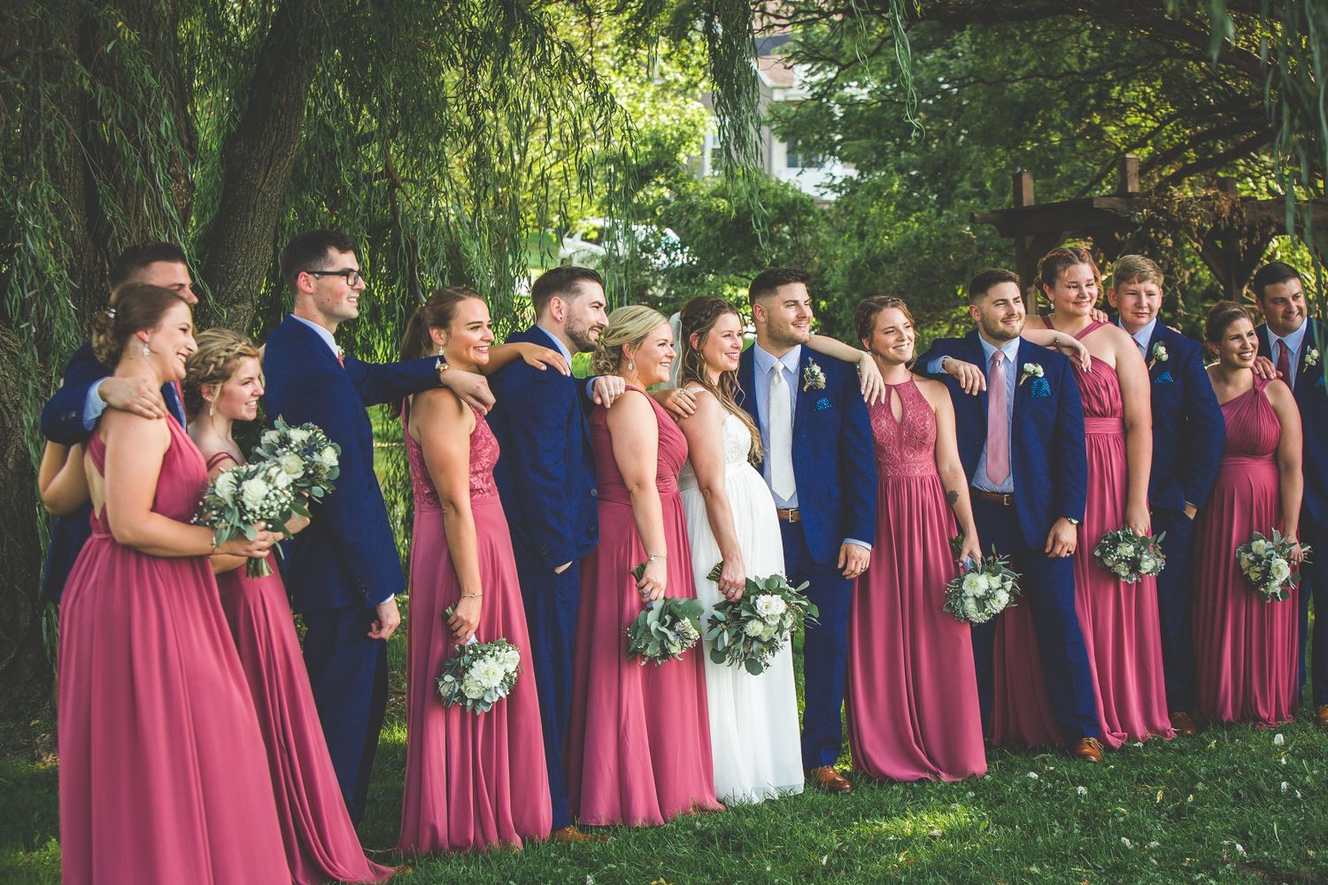 Wedding party in pink bridesmaid dresses and navy suits pose together under trees in an outdoor setting.