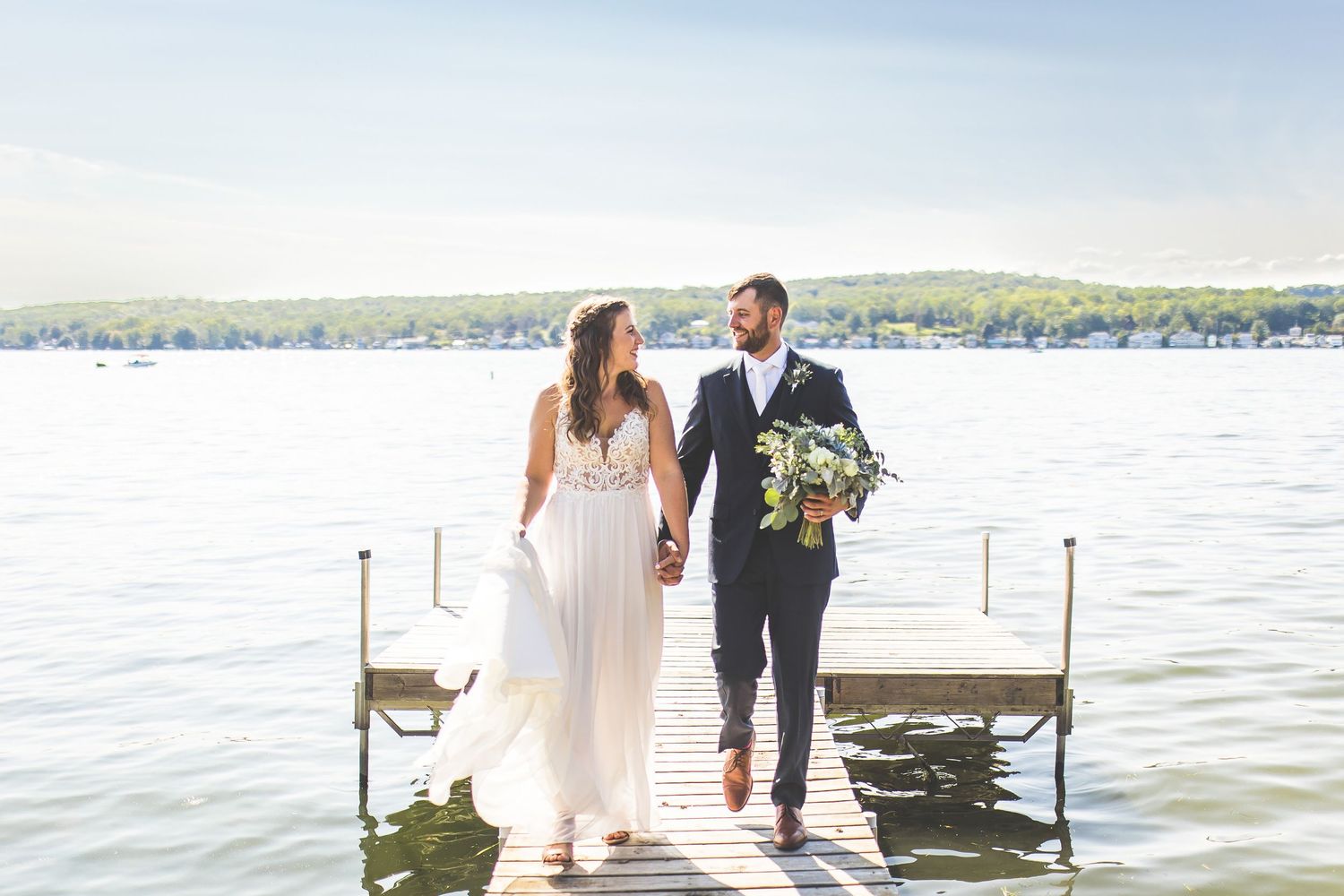 Newlyweds walk hand in hand on wooden dock by scenic lake.