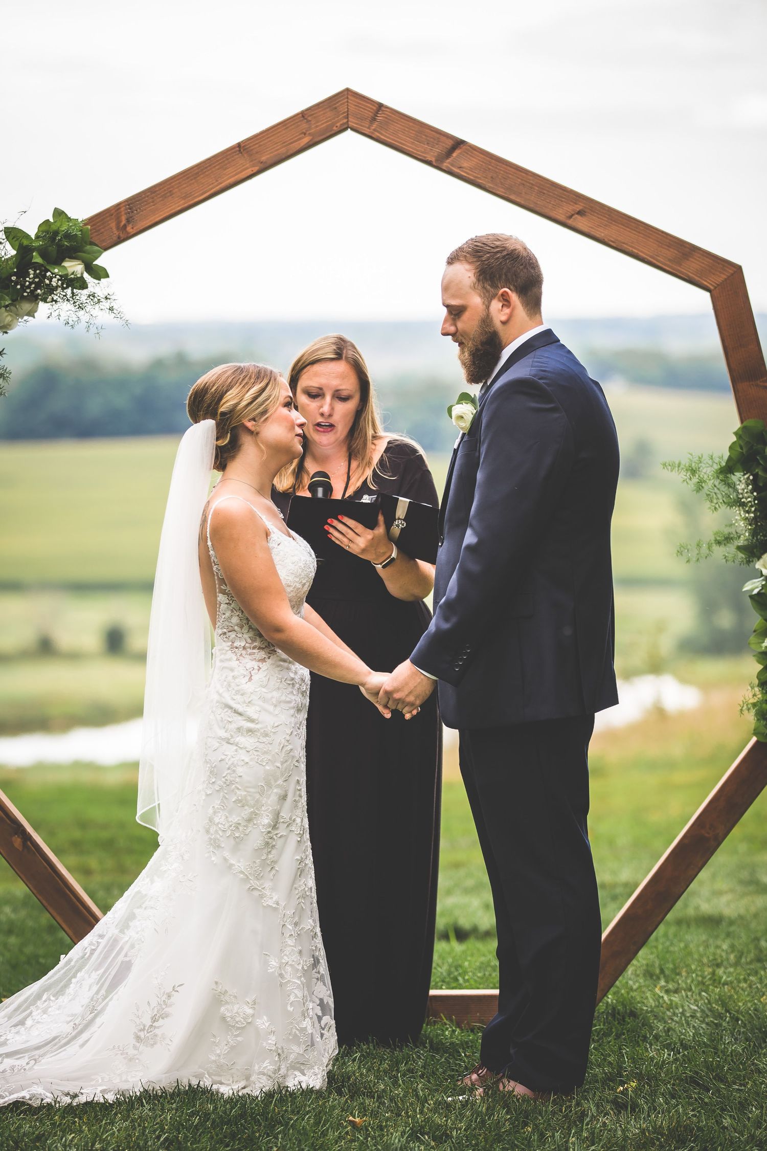 Wedding ceremony under wooden hexagonal arch with rolling hills in background.
