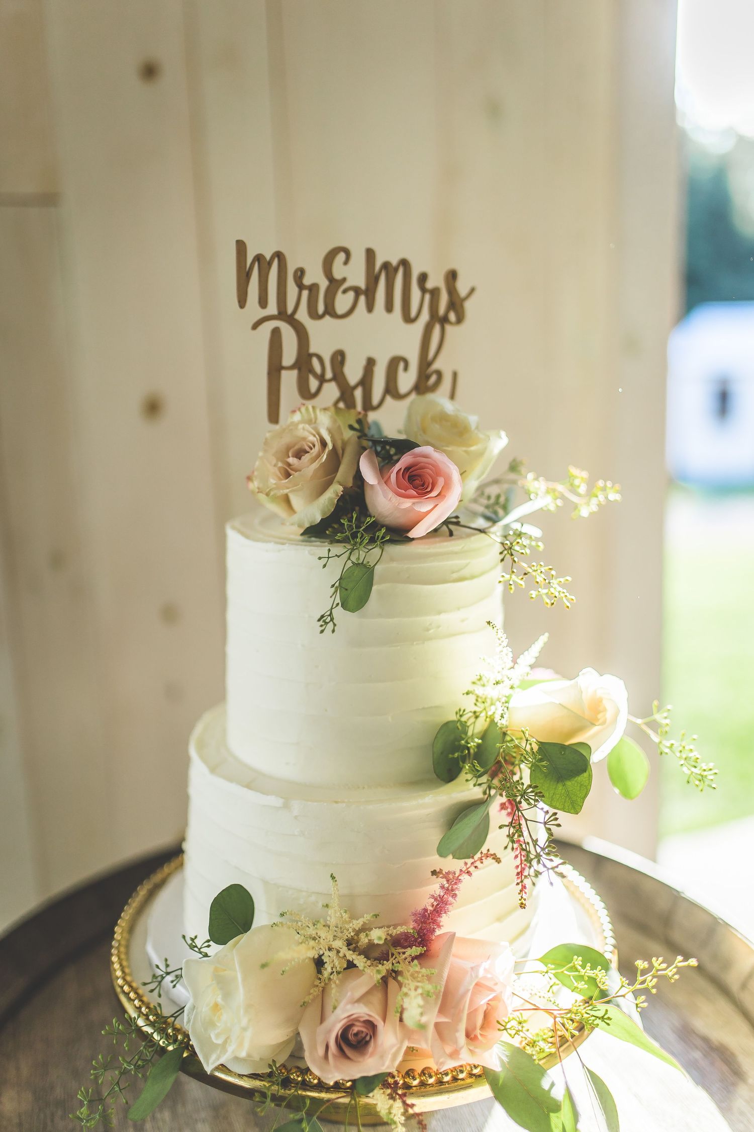 Elegant white wedding cake decorated with pink roses and greenery on wooden stand.
