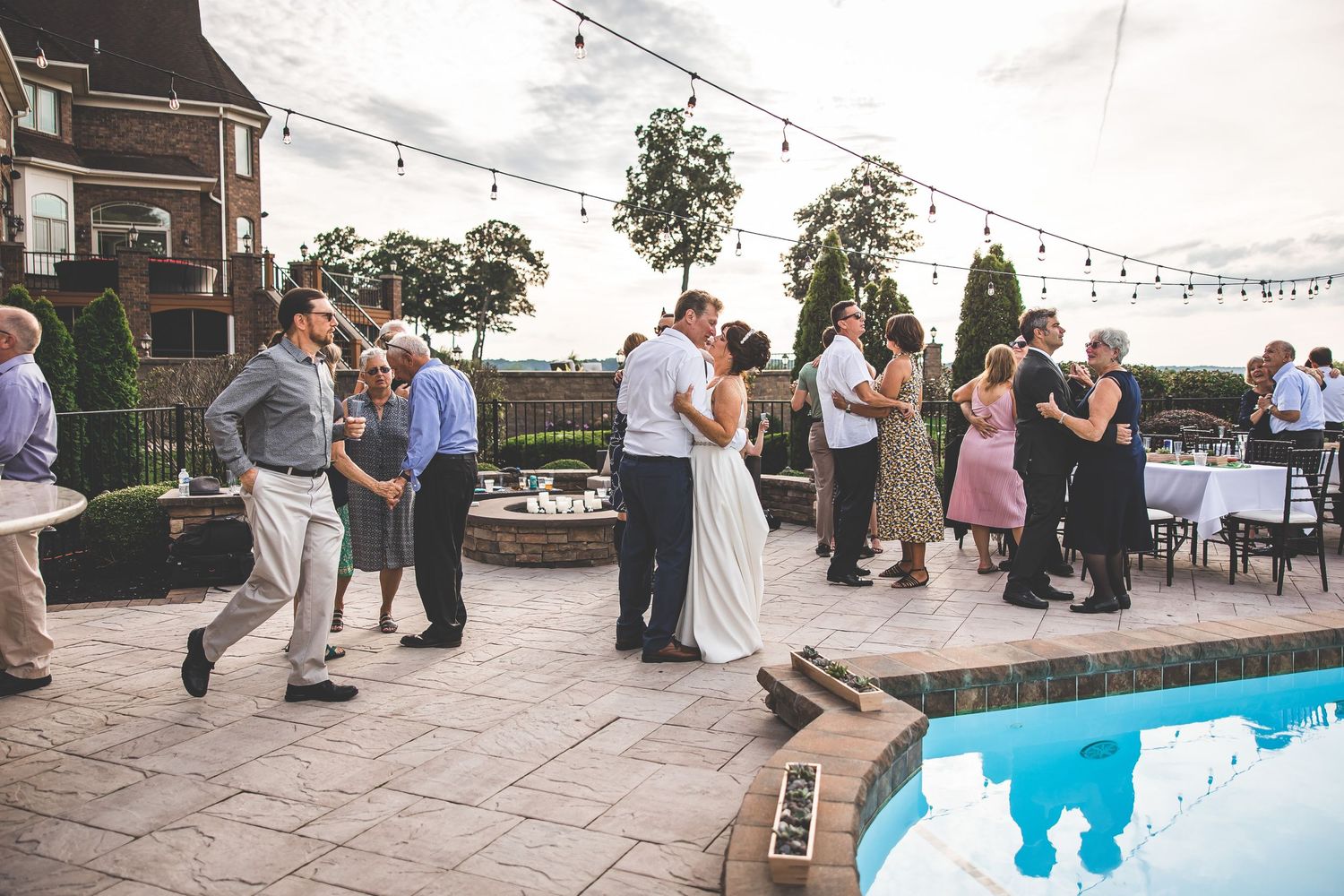 Couples dancing on a stone patio near a swimming pool with string lights overhead.
