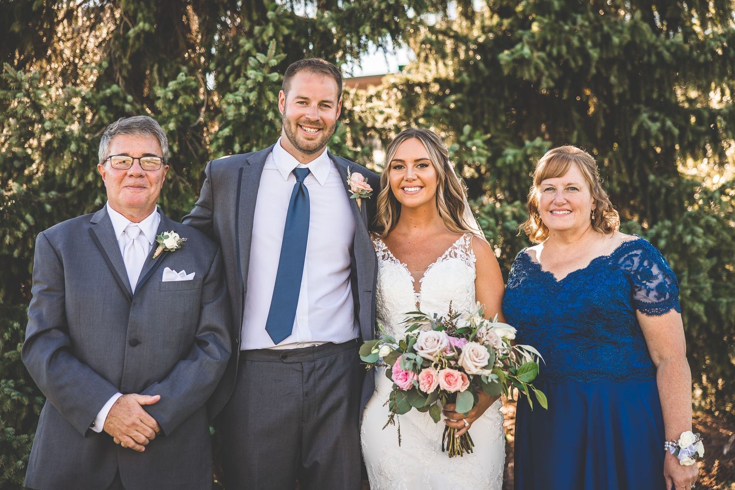 Formal wedding portrait with couple and family members standing in front of evergreen trees.