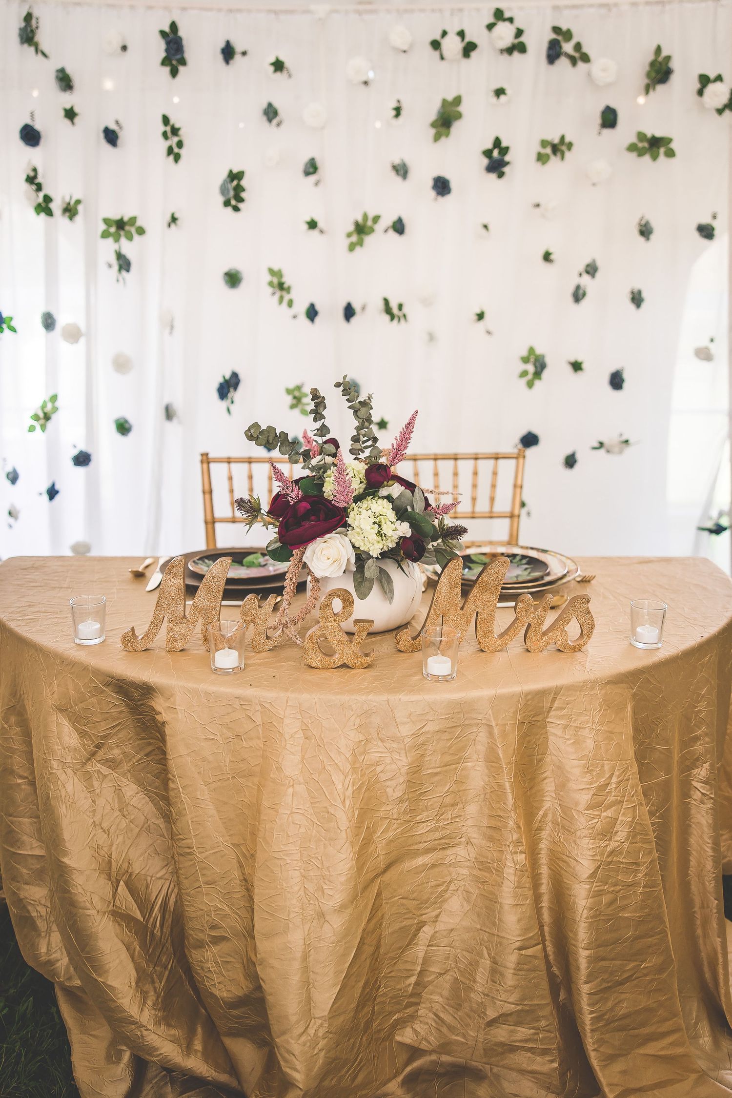 Wedding sweetheart table with gold tablecloth and floral wall backdrop.