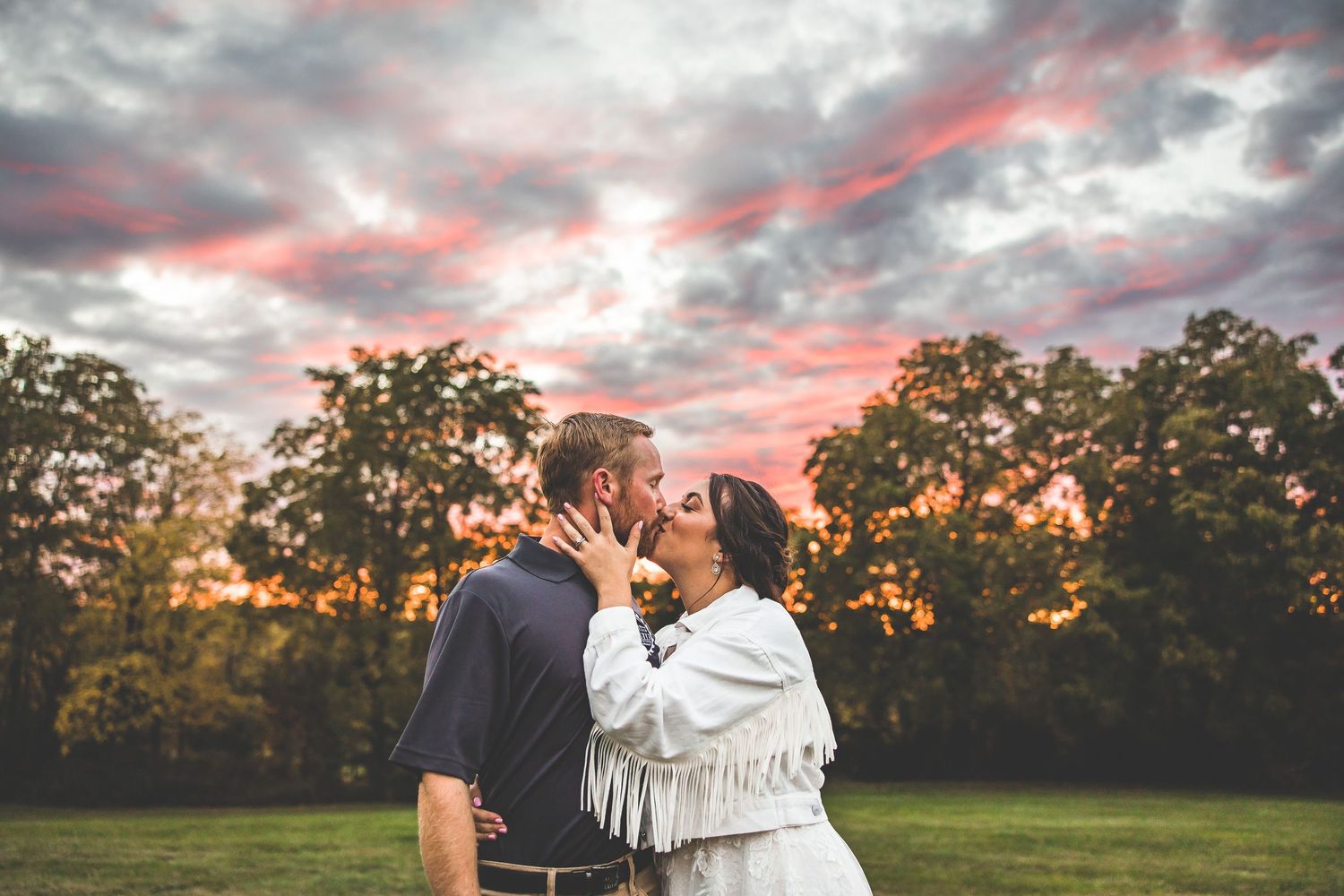 A couple shares a romantic moment at sunset in a grassy field with dramatic pink and orange clouds overhead.