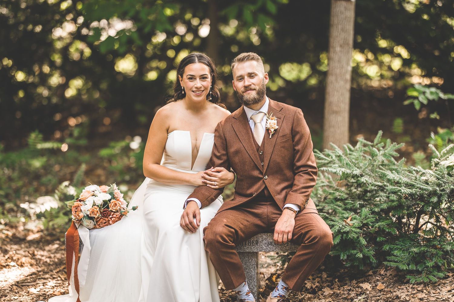 Happy newlyweds laugh together while sitting on fallen logs in a forest setting.