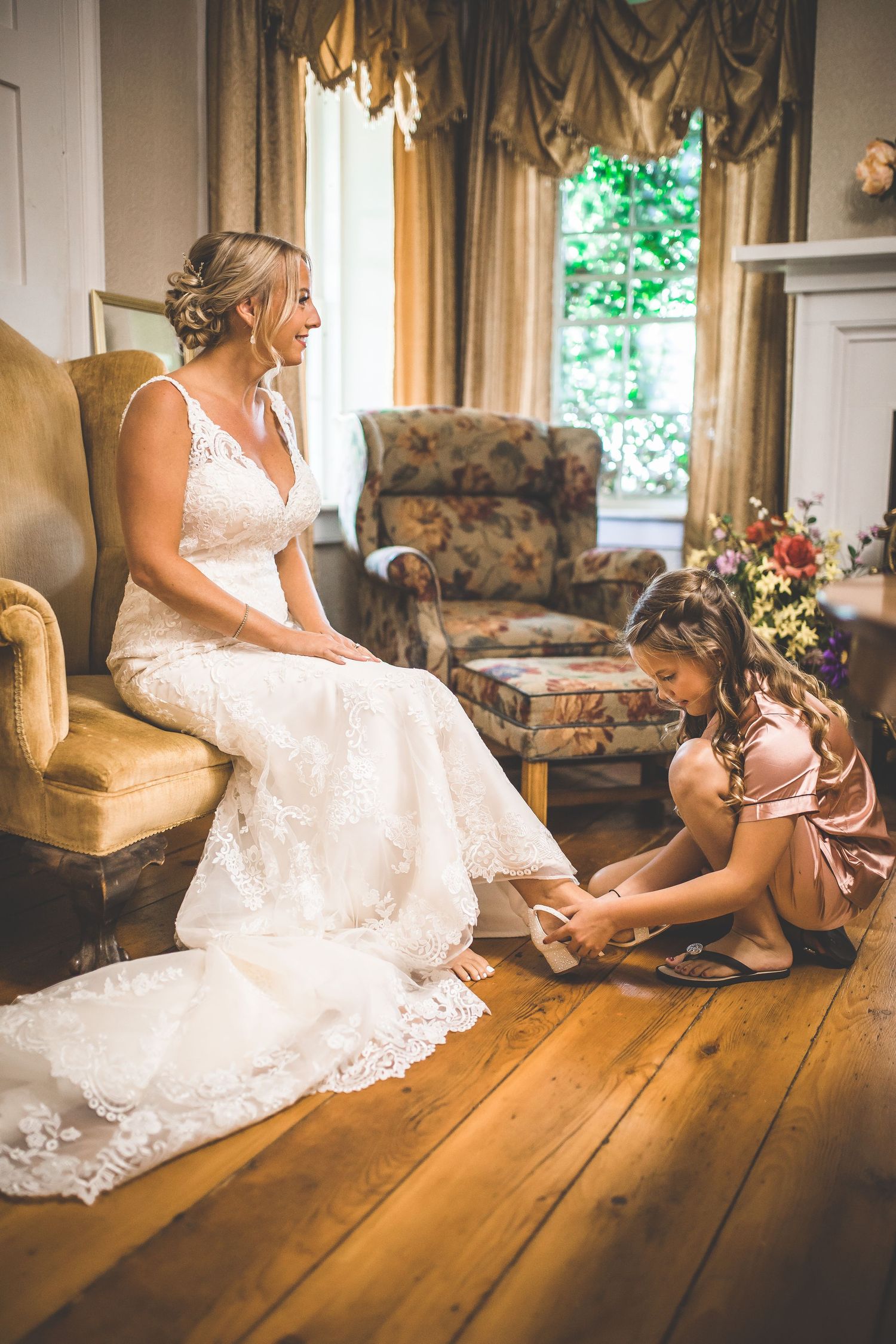 Bride in lace gown seated in vintage chair while final dress preparations are made.