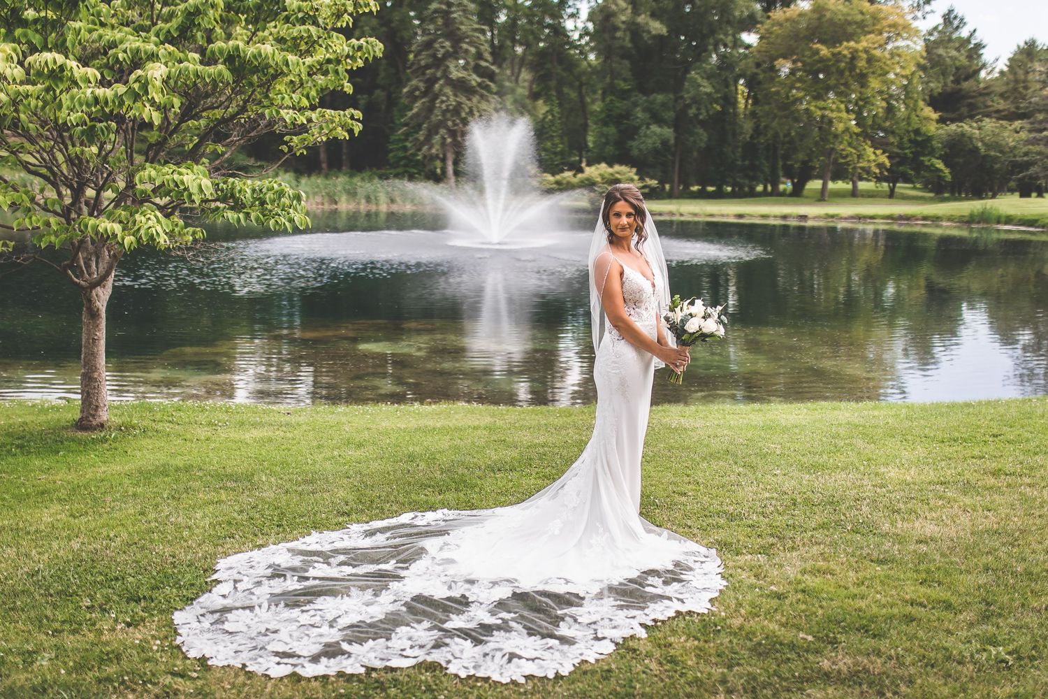 A scenic outdoor wedding photoshoot by a lake with a fountain and lush greenery in the background.