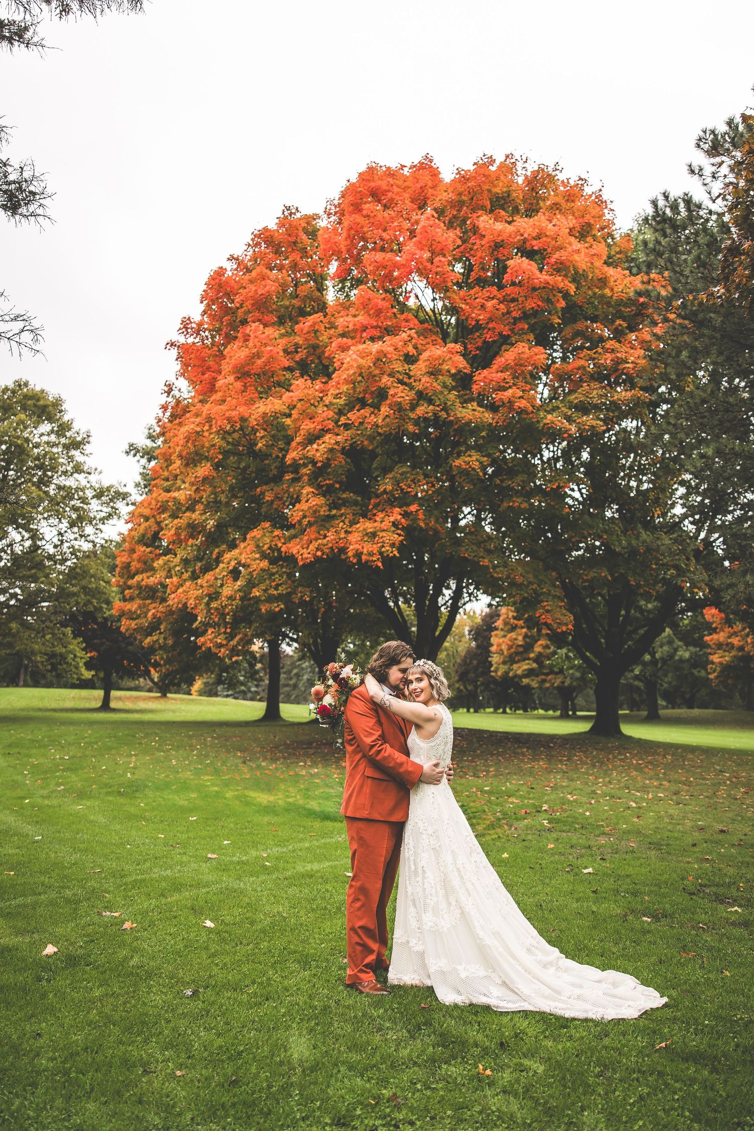 A couple embraces under a vibrant orange autumn tree on a green lawn during their wedding photoshoot.