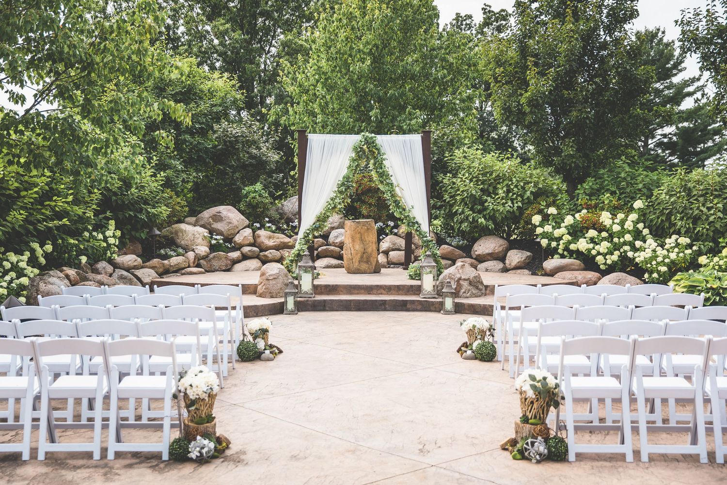 Outdoor wedding ceremony site with white chairs and floral arch set against natural stone backdrop.