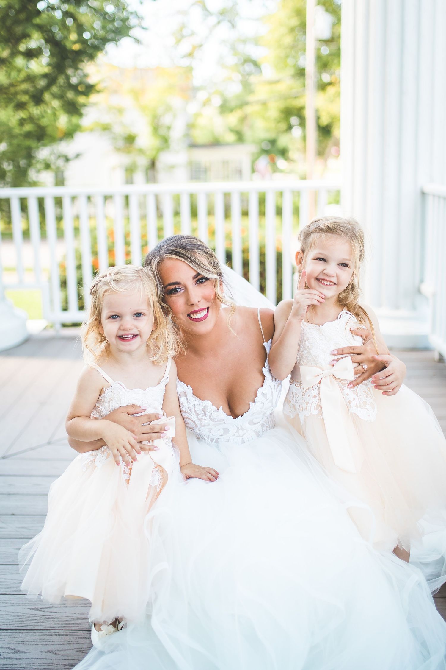 A bride in a white gown poses with two flower girls in tulle dresses on a porch.