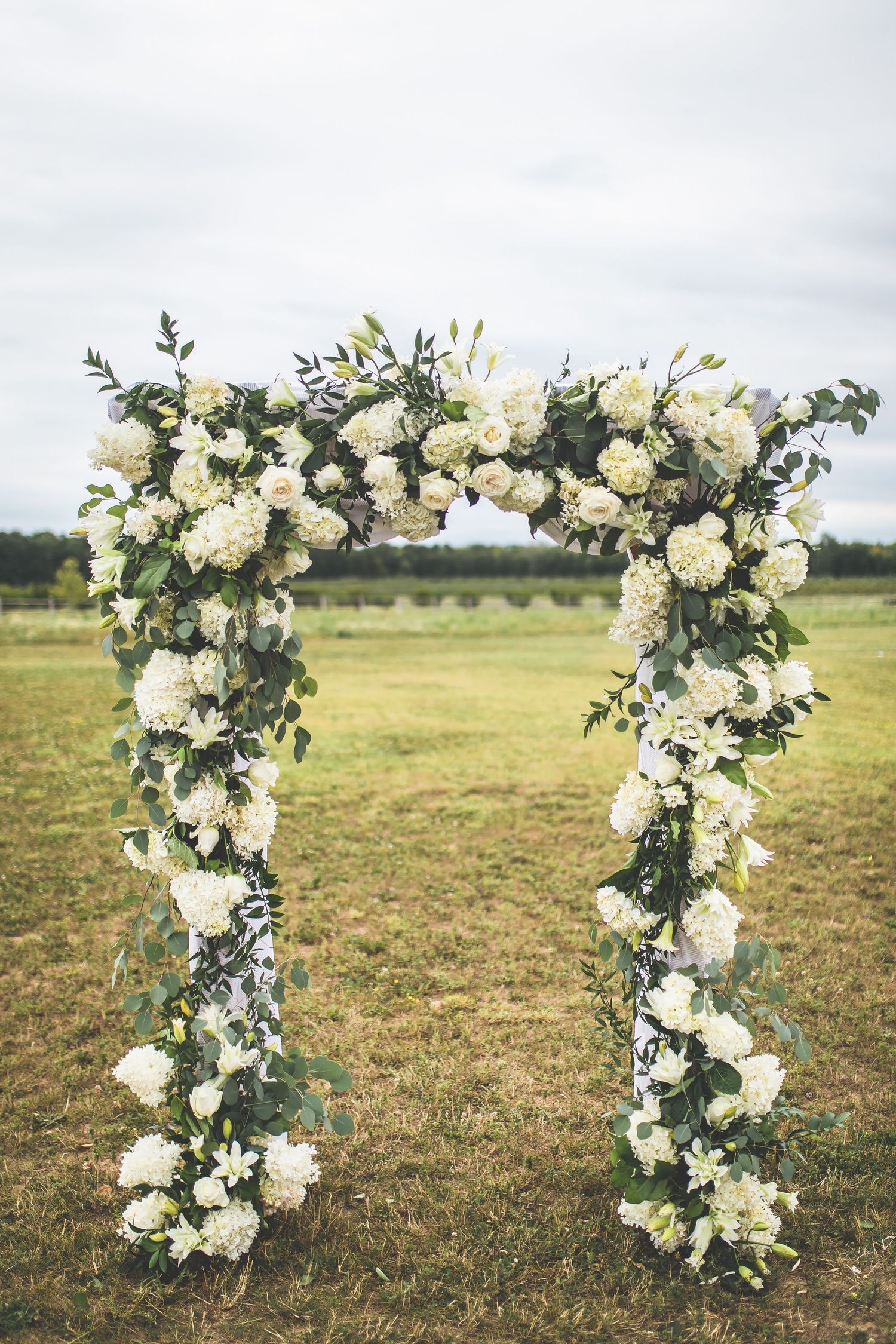 White floral wedding arch with greenery stands in an open field.