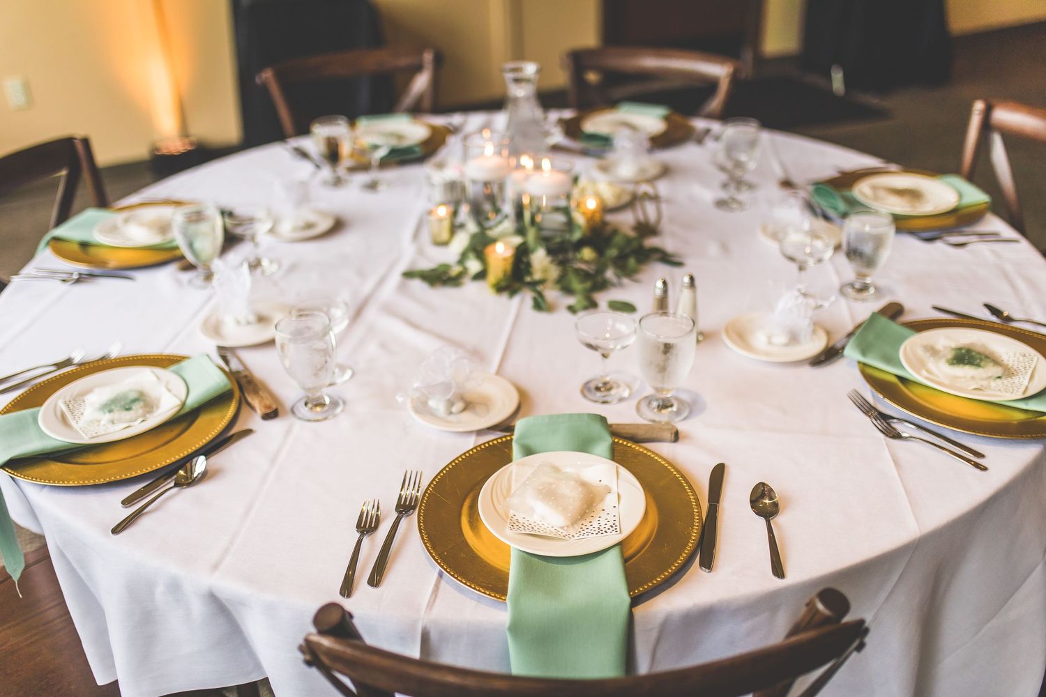 An elegant wedding reception table set with gold charger plates, mint green cups, and white floral centerpieces.