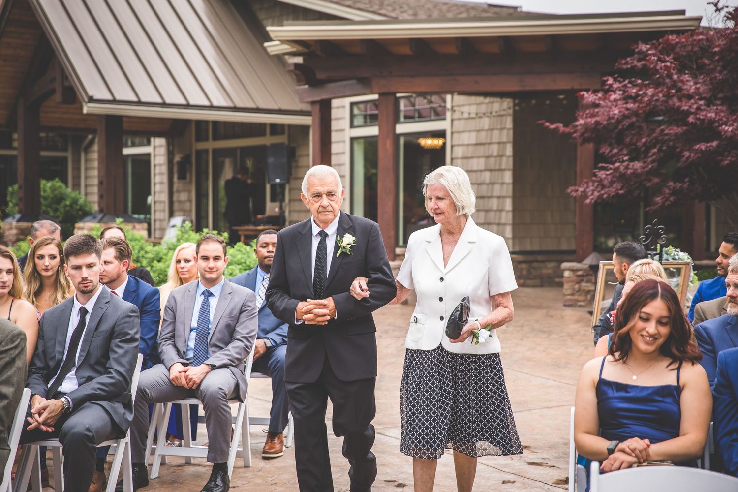 Wedding guests in formal attire gather outdoors at a ceremony venue with brick and stone architecture.