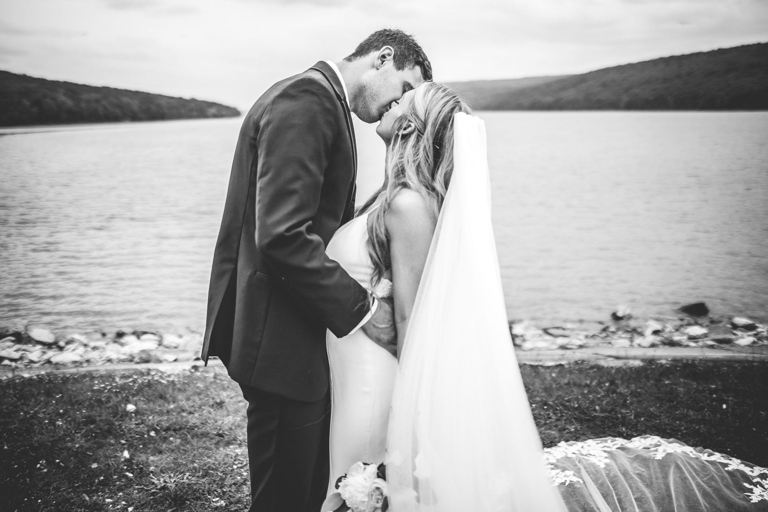 Black and white wedding photos capture intimate moments between newlyweds by a scenic waterfront.