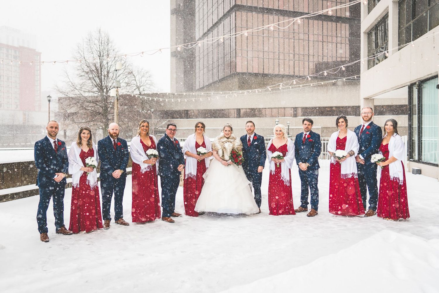 Wedding party stands together in falling snow wearing red bridesmaid dresses and navy suits against modern building.