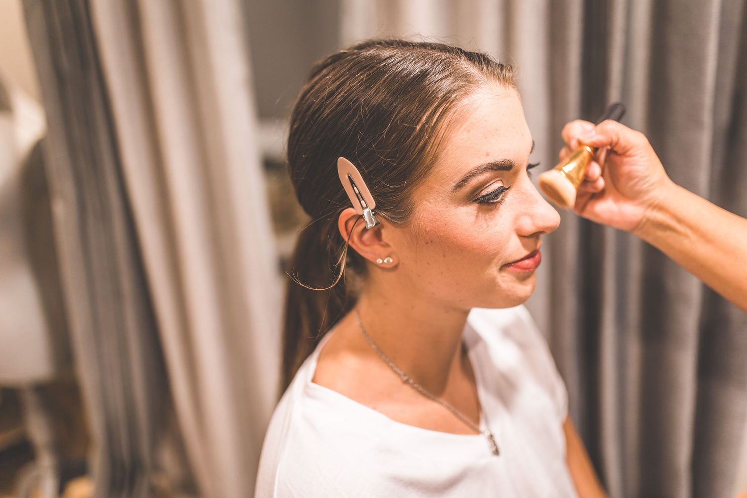 Soft morning light illuminates bridal preparation with elegant curtains in background.