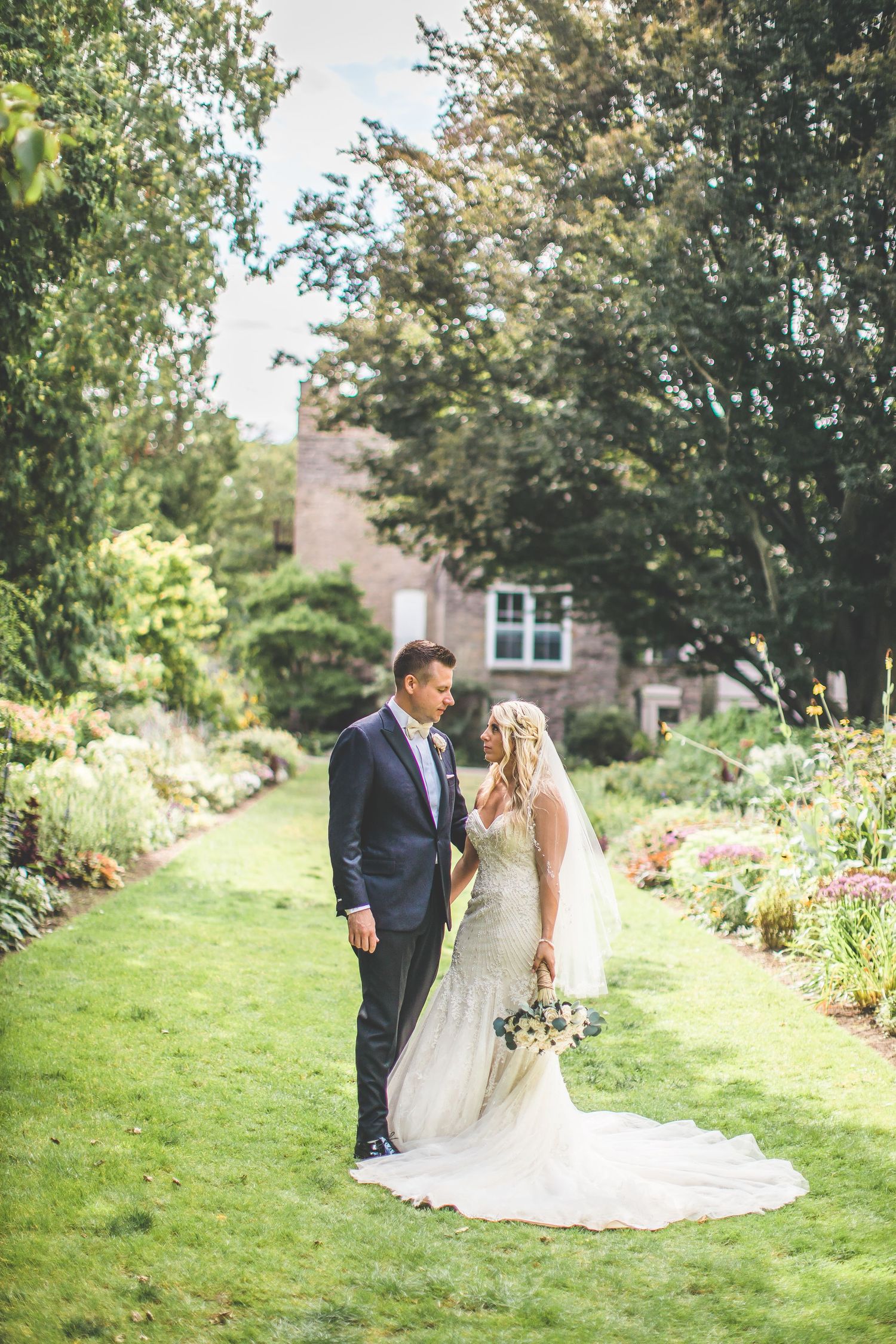 Couple stands together on garden path surrounded by blooming flowers and greenery.