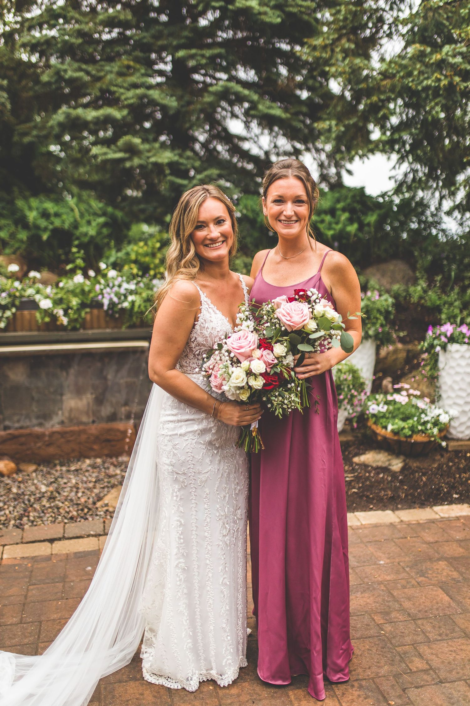 Bride in white lace gown and bridesmaid in dusty rose dress holding floral bouquets on brick patio.