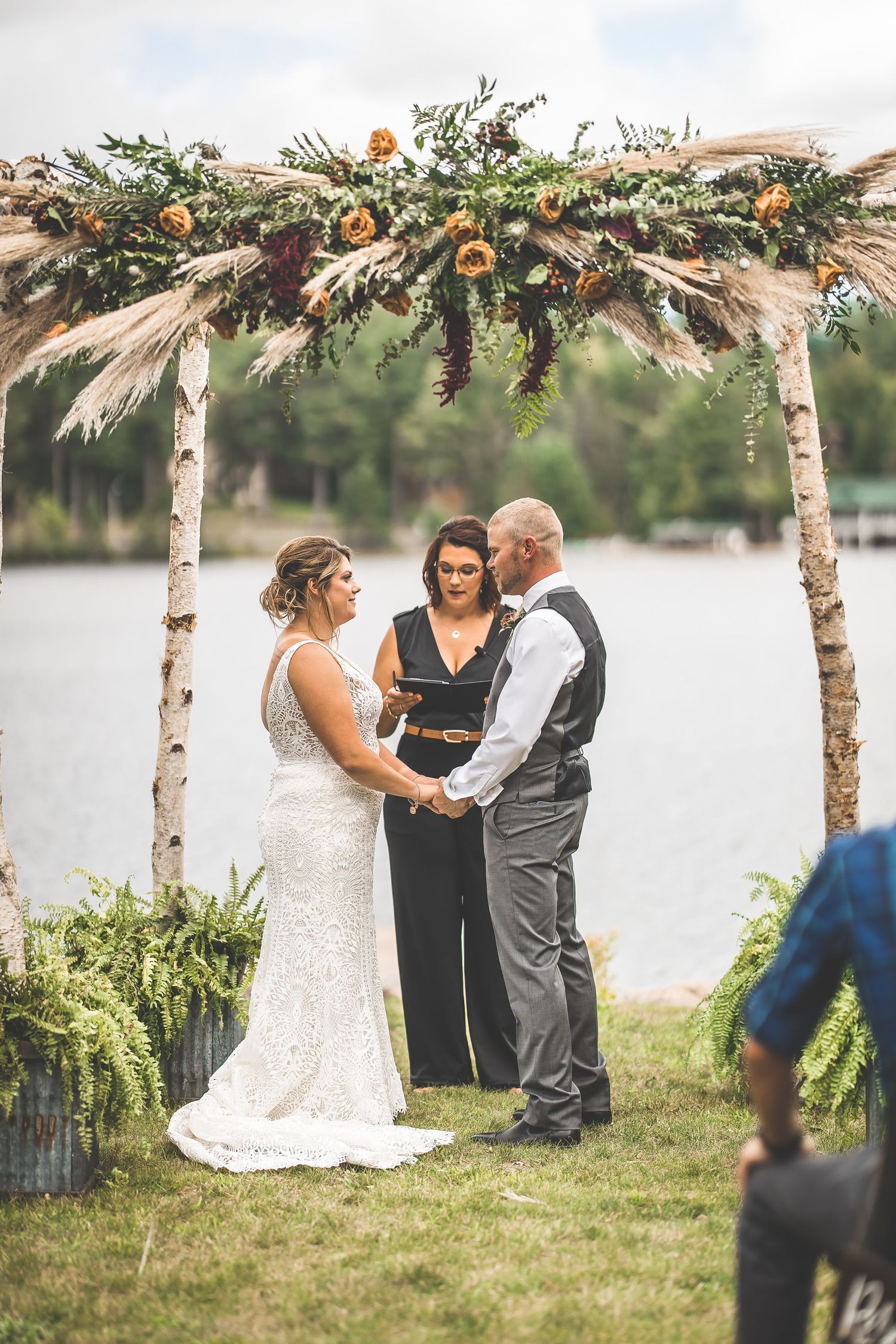 Wedding ceremony under a birch arch adorned with pampas grass, greenery and orange flowers beside a lake.