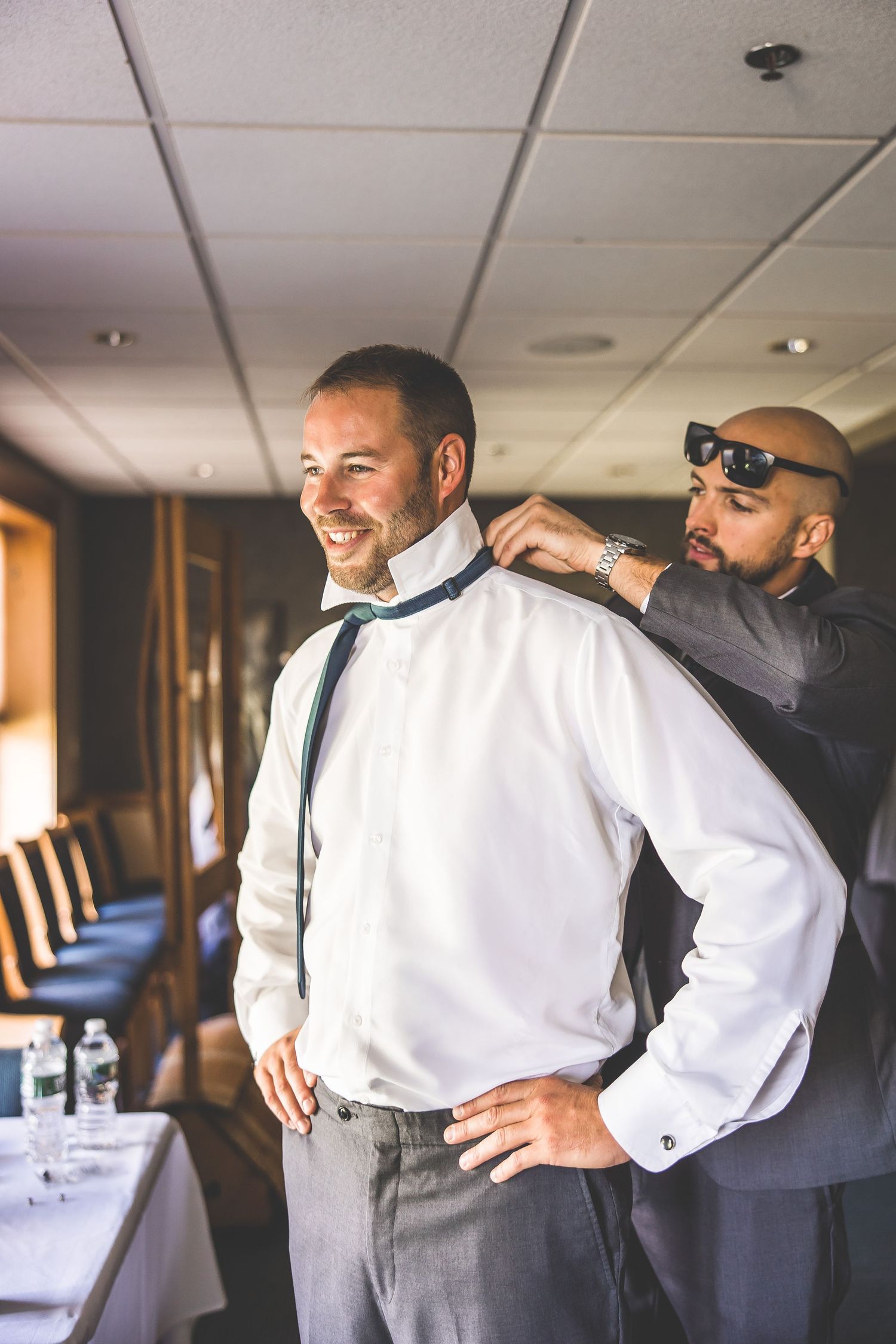 Groomsmen help each other get ready by adjusting ties and suspenders in a formal indoor setting.