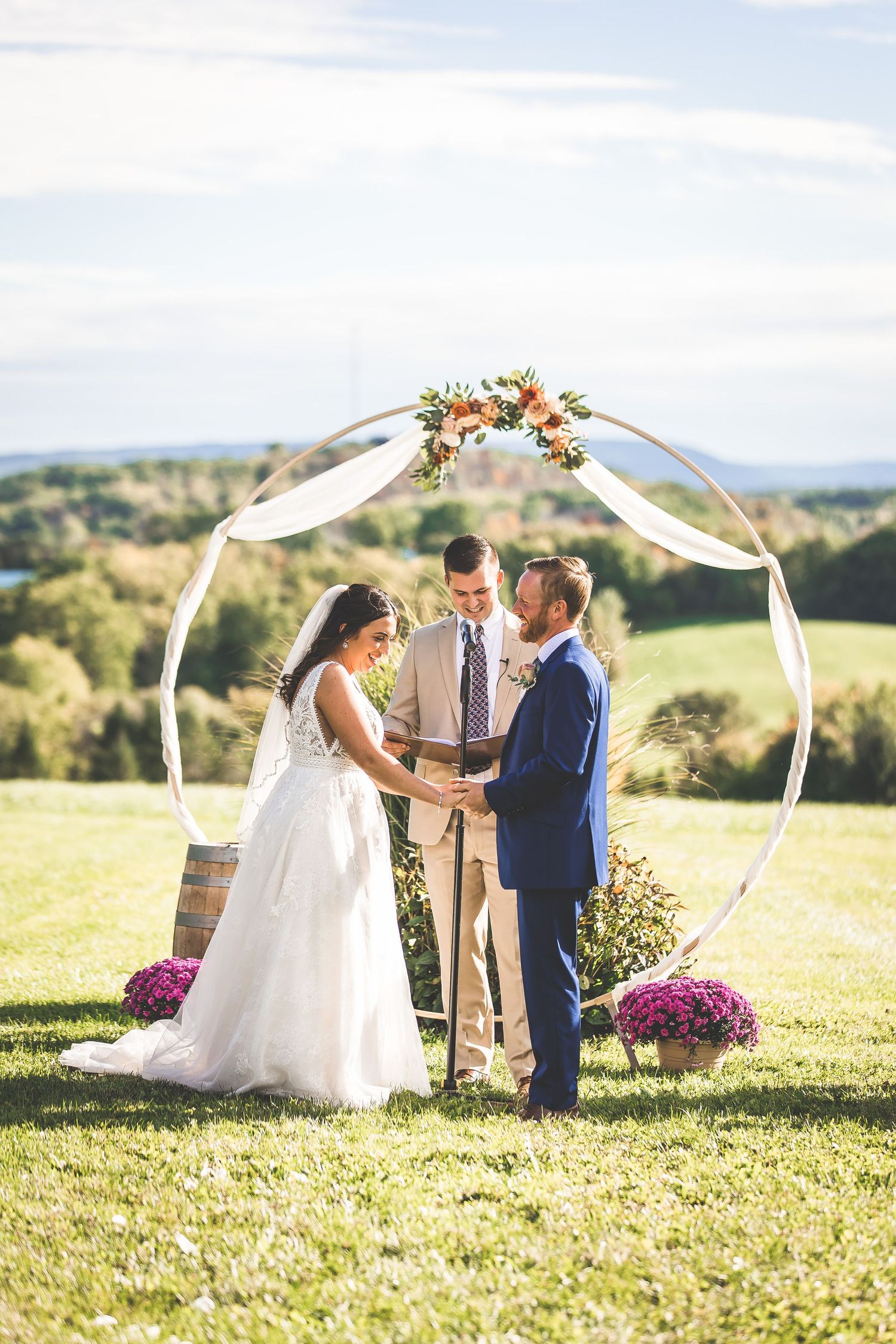 Couple exchanges vows beneath a circular arch adorned with peach flowers and white fabric on a scenic hilltop.
