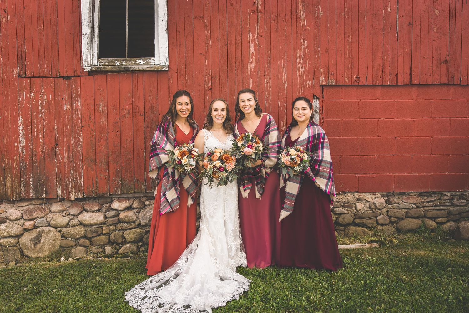 Wedding party poses in front of a weathered red barn on a sunny day.