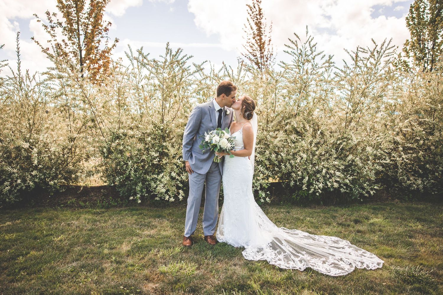 Bride and groom share tender moment in sunlit orchard setting.