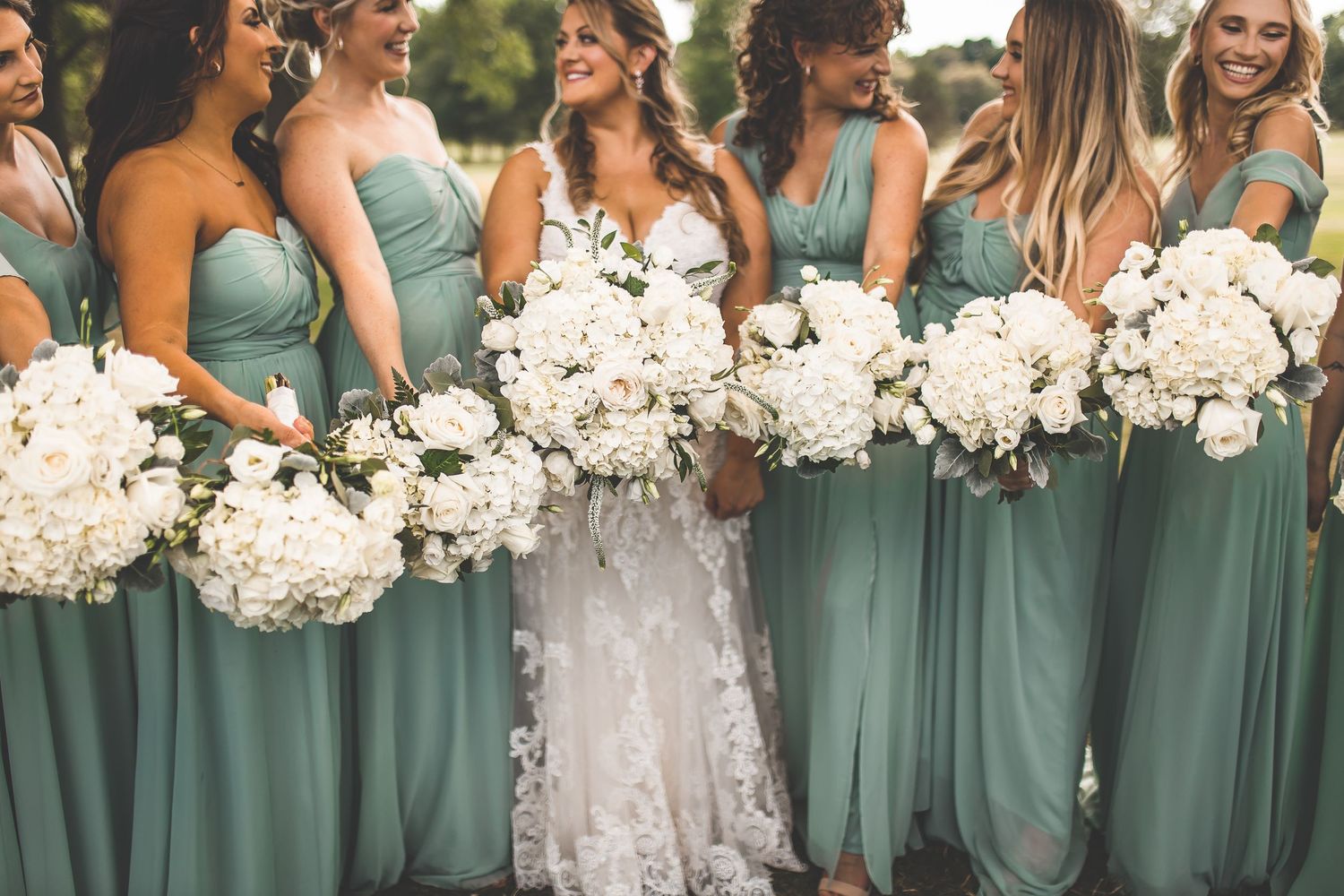 Bridesmaids in sage green dresses hold white bouquets while standing in a row outdoors.