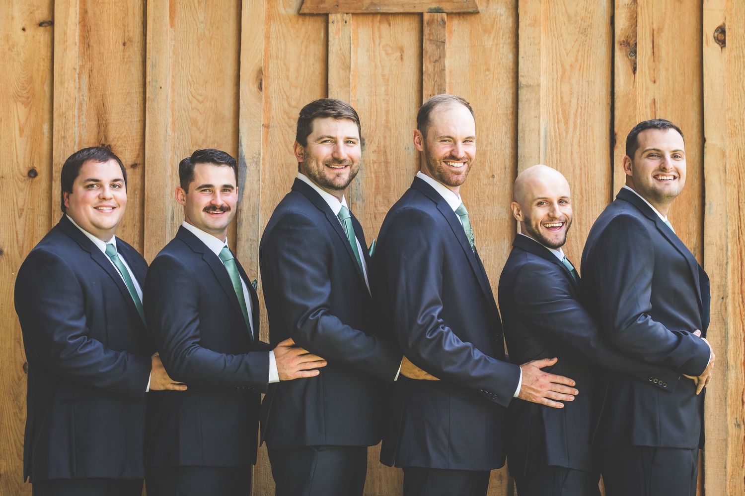 Groomsmen in navy suits pose playfully in a line against a wooden wall backdrop.