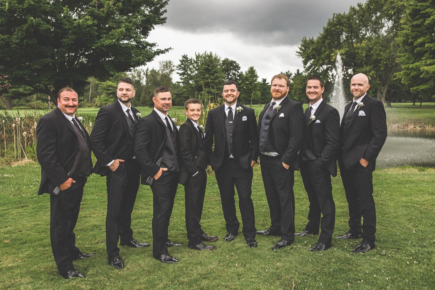Groomsmen in black suits posing together outdoors on a cloudy day.