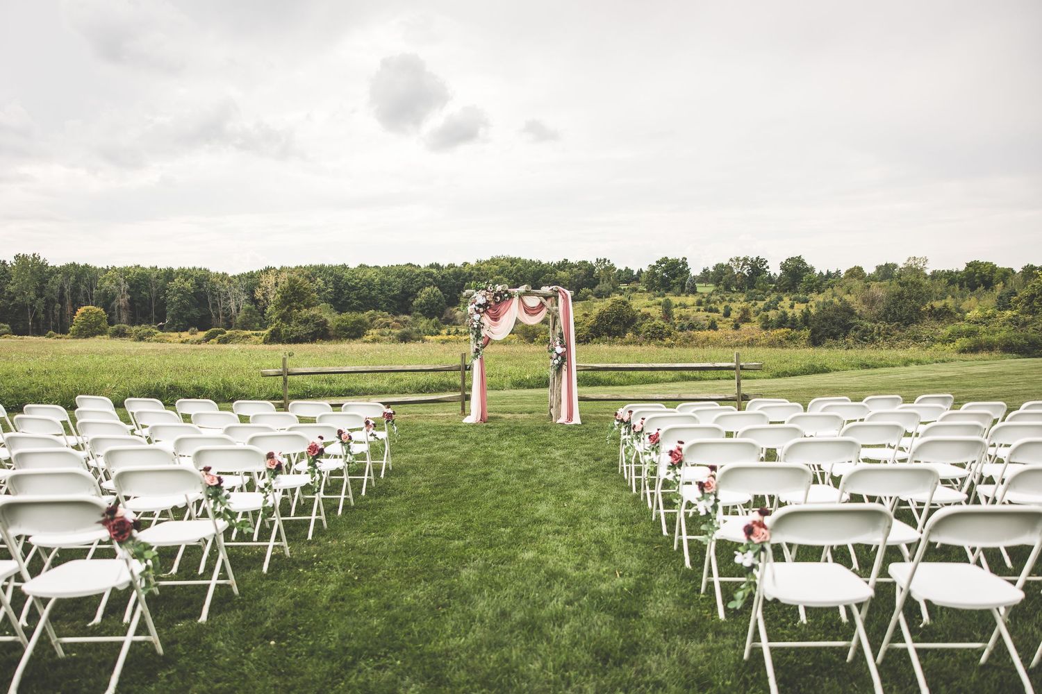 Outdoor wedding ceremony setup with white folding chairs and floral arch on a green lawn.